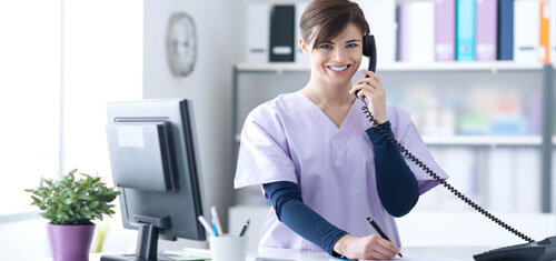 Picture of a receptionist representing Top Plastic Surgeons in beautiful San José, Costa Rica.  The woman has short brown hair, is wearing a hospital smock and is standing at the receptionist desk while smiling at the camera.