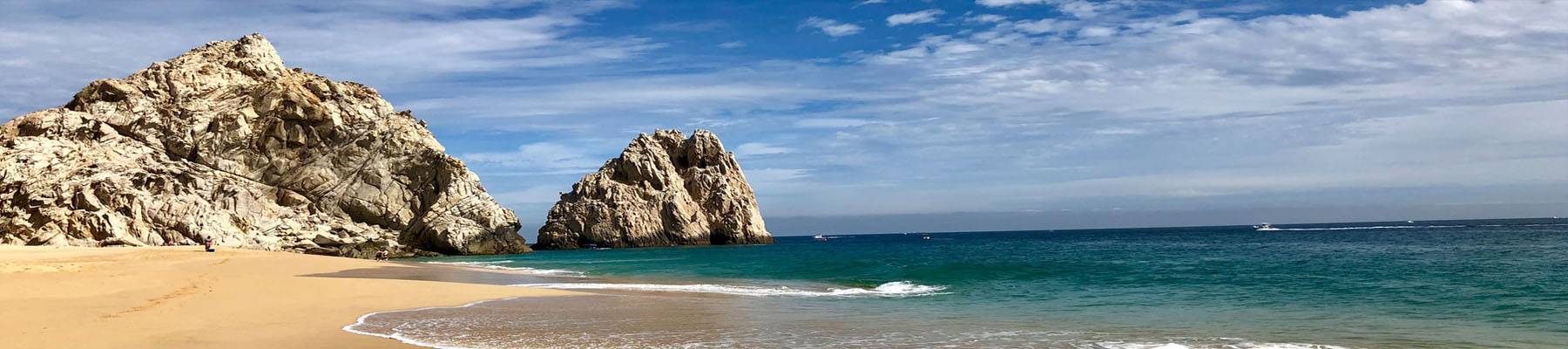 Picture of a beautiful beach in Jaco.  The picture shows tall rocks, a sandy beach, blue water and a blue sky with white clouds.