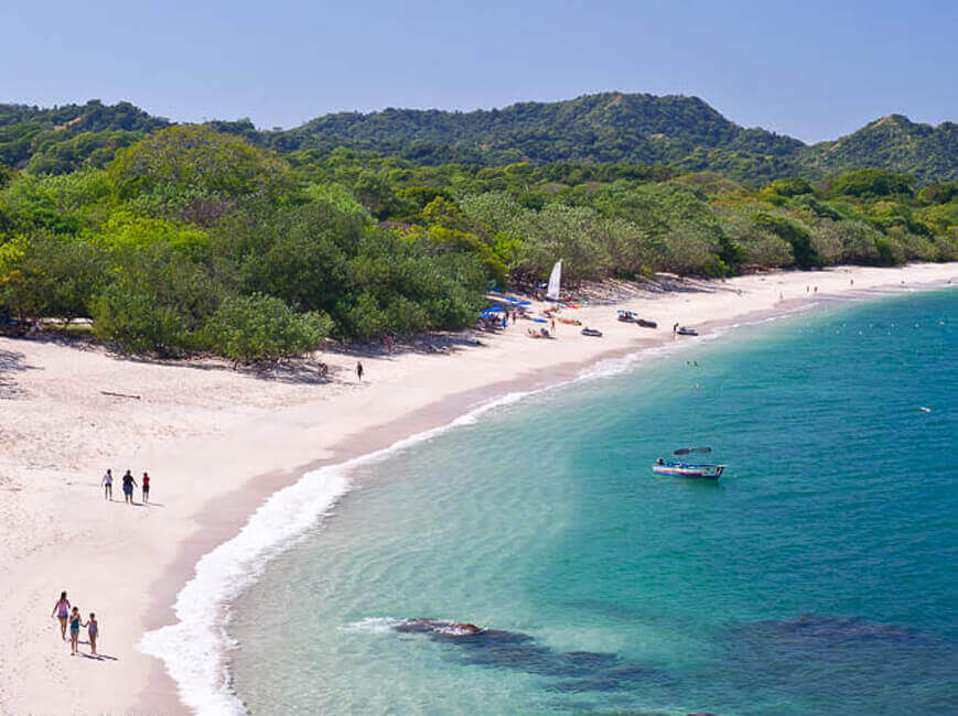 Picture of a beautiful sandy beach in Costa Ricas.  The picture shows white sand, blue water and framed by tall rocks behind the shoreline.