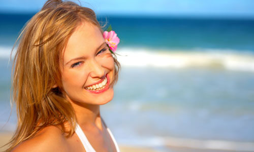 Close-up picture of a beautiful woman, happy with her face lift with neck lift in San José, Costa Rica.  The woman has long sandy blonde hair and is looking directly at the camera with the ocean behind her.