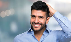 Picture of a man, happy with his hair transplant procedure he had with Top Plastic Surgeons in beautiful San José, Costa Rica.  The man is wearing a blue shirt with an open collar and holding his left hand to his head.