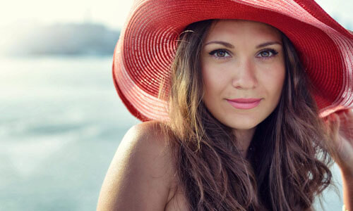 Portrait picture of a beautiful woman, happy with her face lift with neck lift procedure in San José, Costa Rica.  The woman has long dark hair, is looking directly into the camera and is wearing a soft red beach hat highlighting her face lift with neck lift.